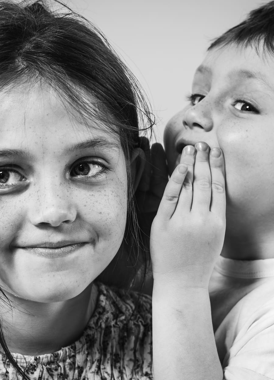 Geschwisterfotografie: Ein Mädchen flüstert einem anderen Mädchen etwas ins Ohr